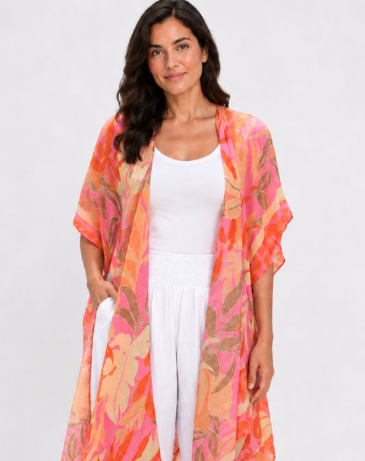 A woman modeling a lightweight, open-front kimono cardigan with a vibrant floral and leaf pattern in shades of orange, pink, and beige, worn over a white tank top and white pants against a light gray studio background.