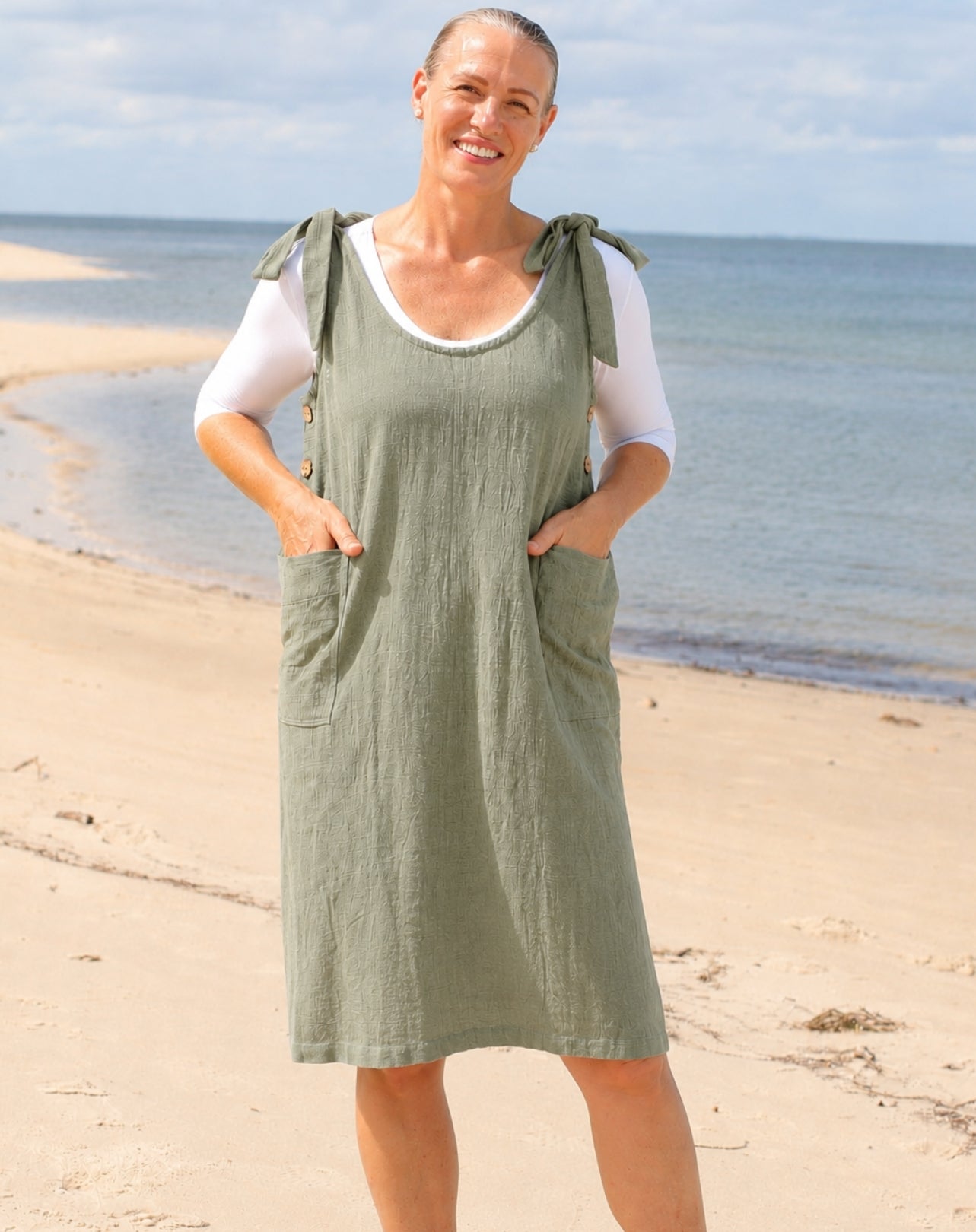A woman standing on a sandy beach wearing an sage green linen-look pinafore dress over a white short-sleeve top.