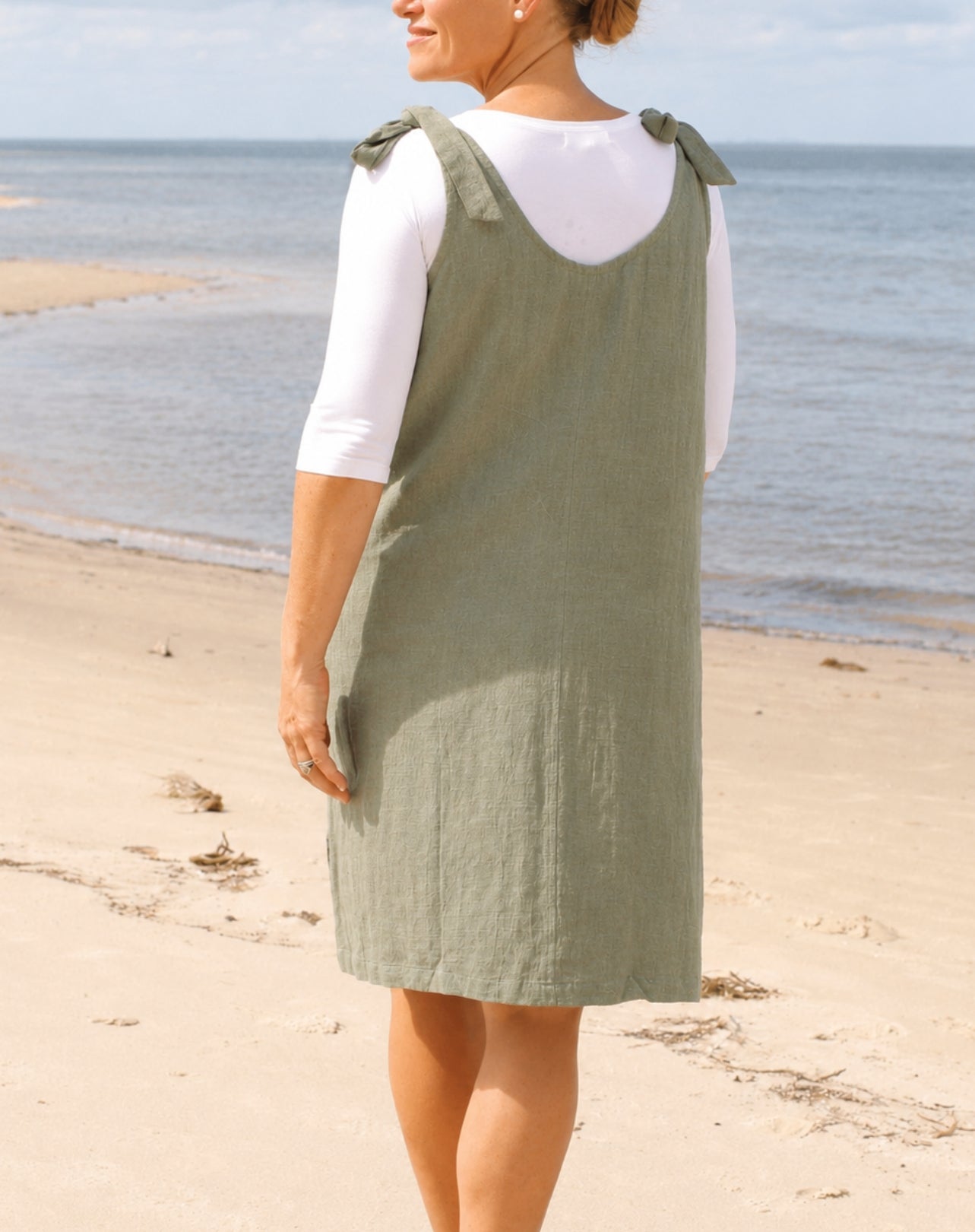A woman standing on a sandy beach wearing an sage green linen pinafore dress over a white short-sleeve top, photographed from behind.