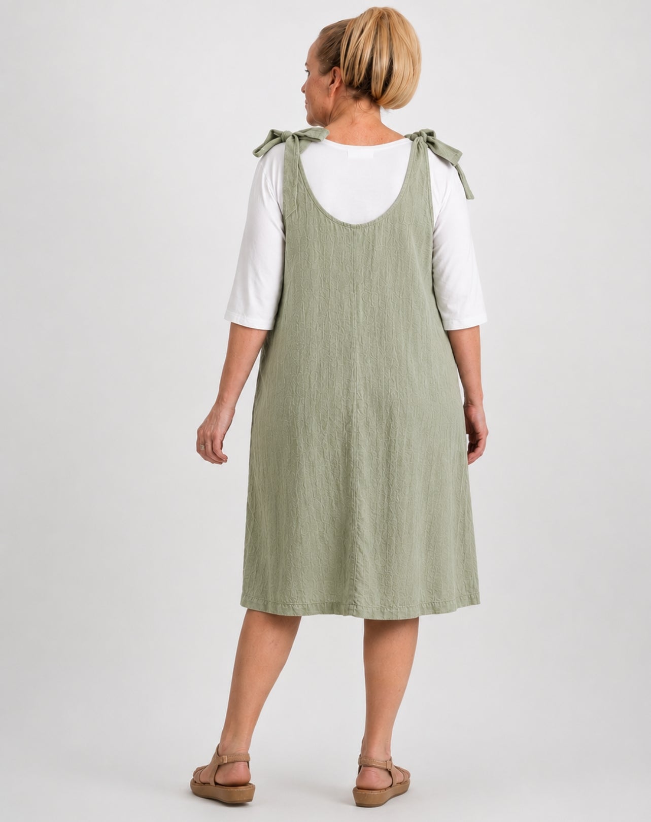A woman shown from the back wearing a sage green sleeveless pinafore-style dress over a white short-sleeve top, paired with brown sandals, against a light gray studio background.