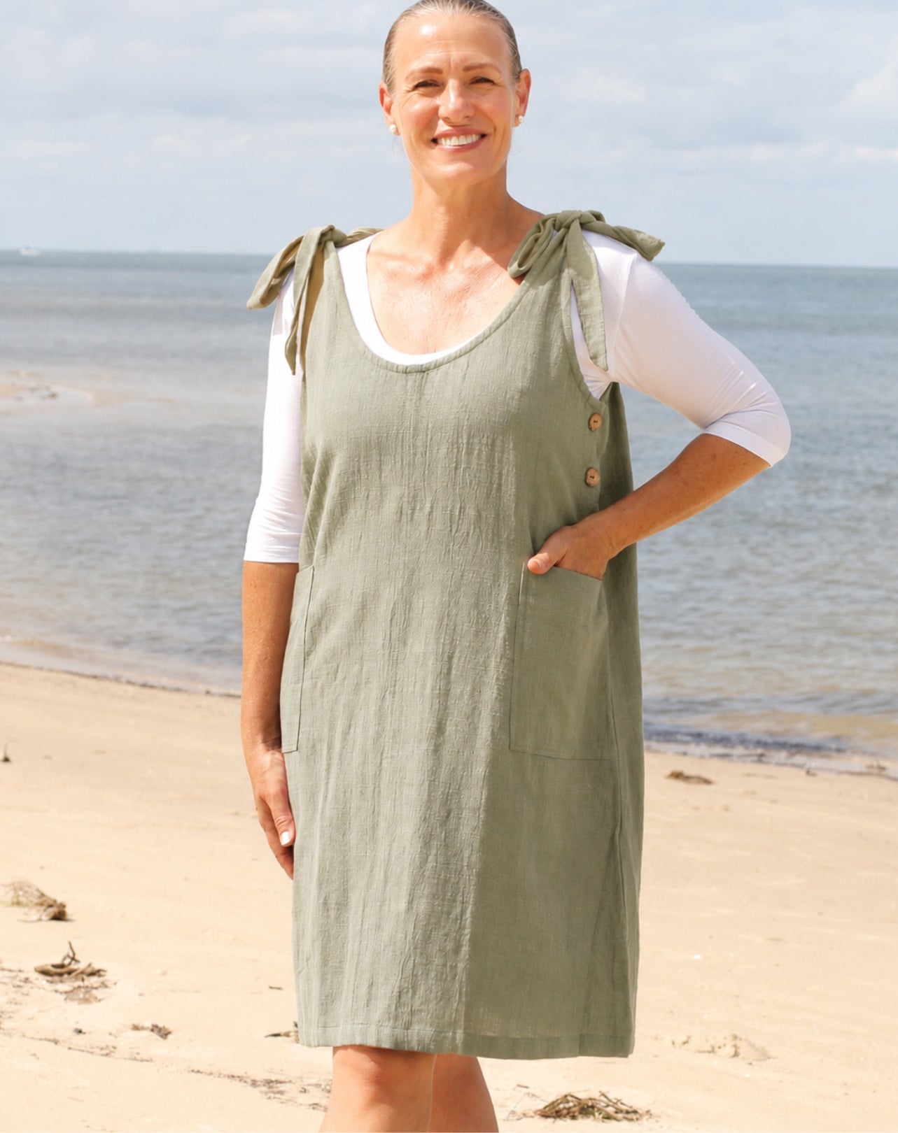 A woman standing on a sandy beach wearing an sage green linen smock dress over a white long-sleeve top. The dress features tie details on the shoulders, a buttoned neckline, and front pockets.