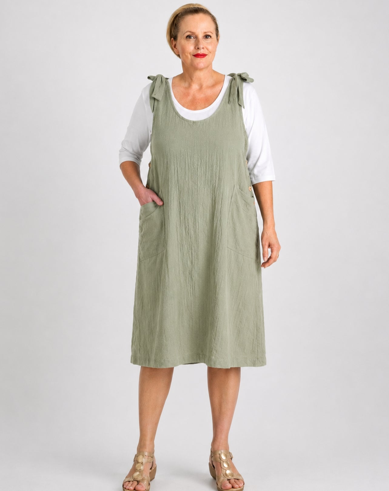 A woman modeling a sage green linen pinafore dress over a white t-shirt, standing against a light gray studio background.