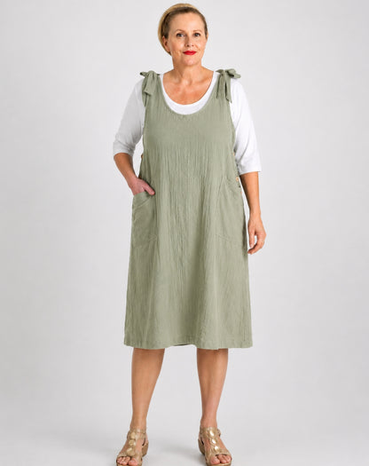 A woman modeling a sage green linen pinafore dress over a white t-shirt, standing against a light gray studio background.