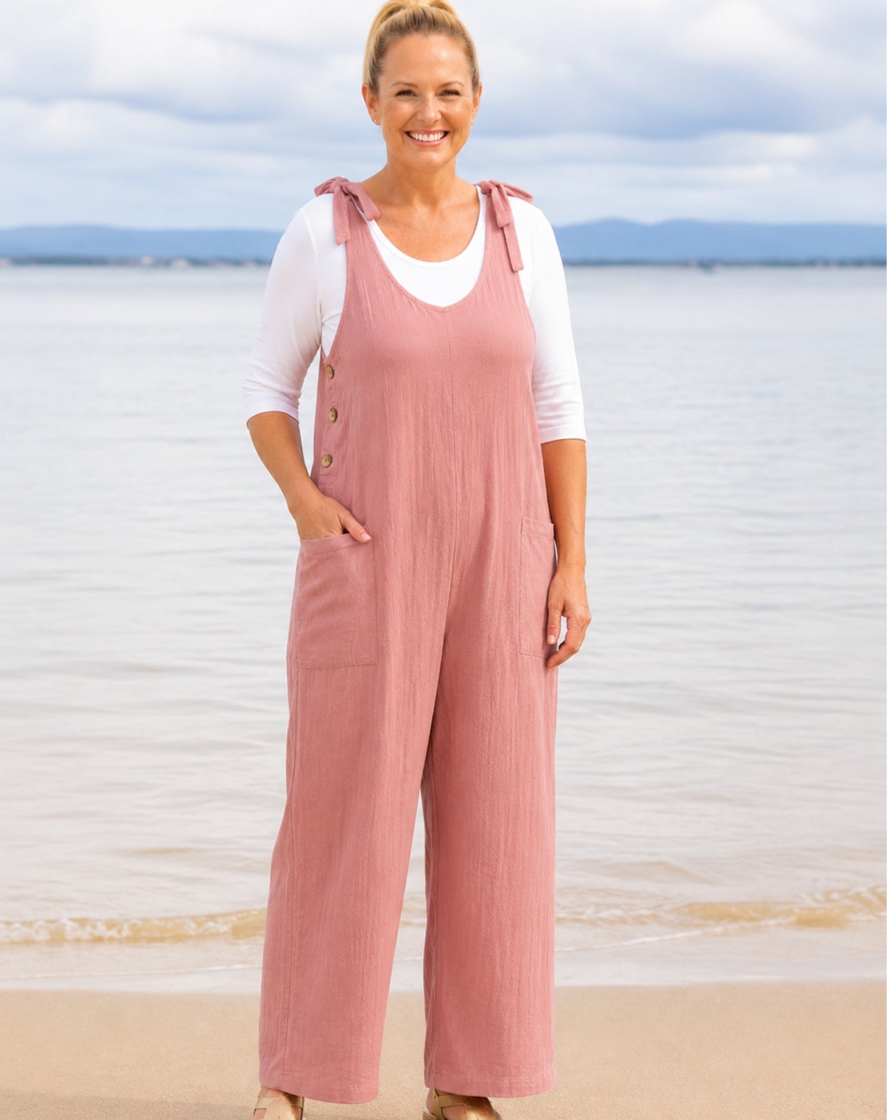 A woman standing on a beach wearing dusty pink wide-leg overall jumpsuit with adjustable tie straps over a white long-sleeve top, with water and cloudy sky in the background.