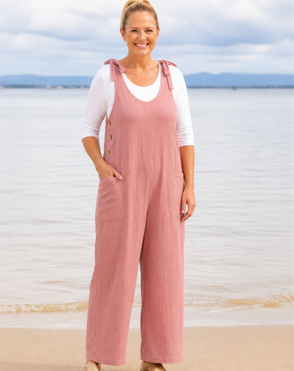 A woman standing on a beach wearing dusty pink wide-leg overall jumpsuit with adjustable tie straps over a white long-sleeve top, with water and cloudy sky in the background.