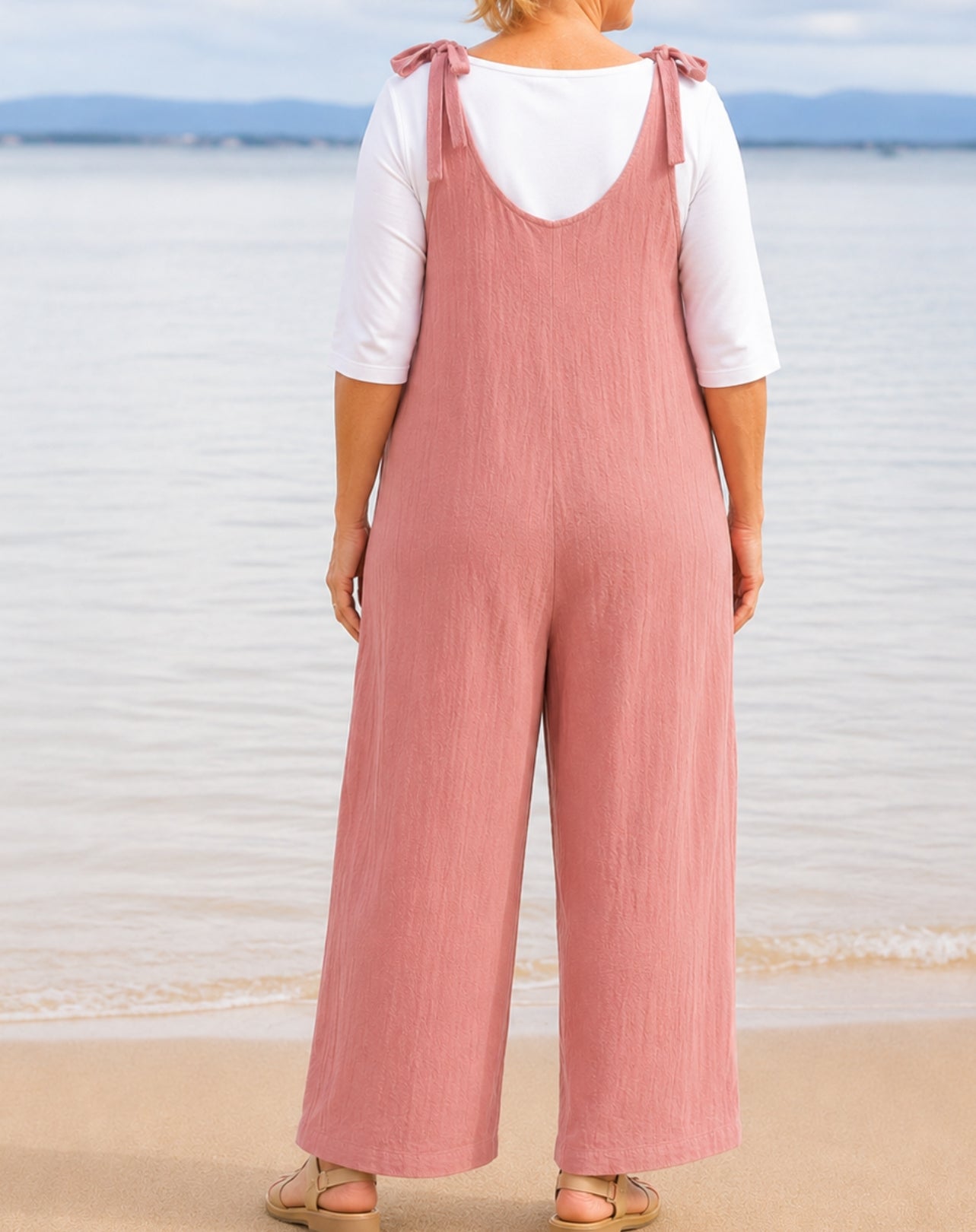 A woman standing on a beach wearing a blushed pink wide-leg jumpsuit with shoulder straps over a white short-sleeve top, shown from the back.