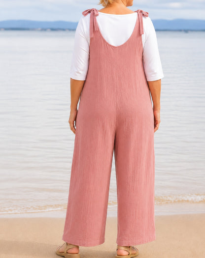 A woman standing on a beach wearing a blushed pink wide-leg jumpsuit with shoulder straps over a white short-sleeve top, shown from the back.