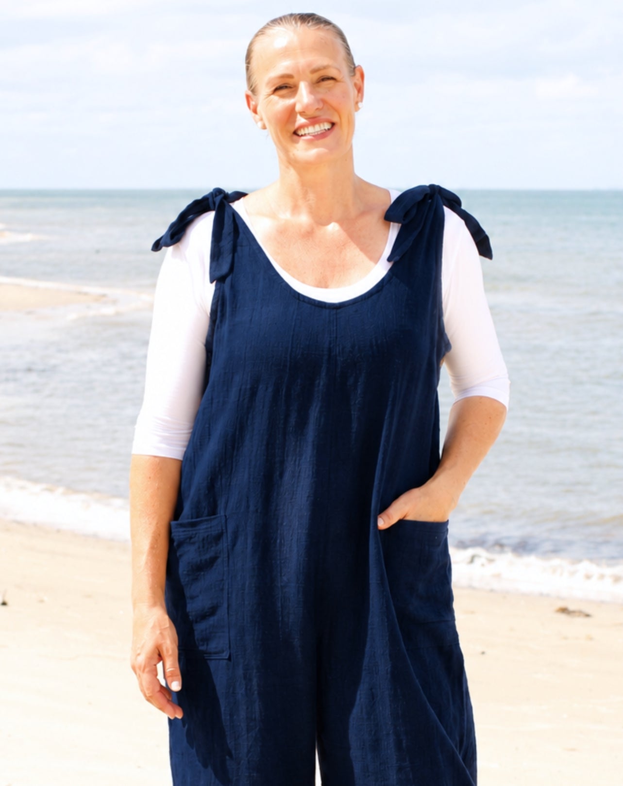 A woman standing on a sandy beach wearing a navy blue linen-look overall jumpsuit with tie shoulder straps over a white short-sleeve top.