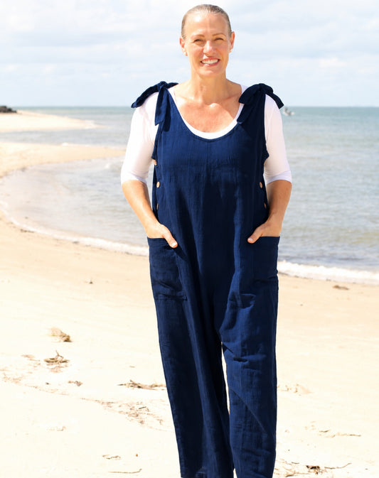 A woman standing on a sandy beach wearing navy blue linen-look overall jumpsuit with shoulder ties, layered over a white short-sleeve top.