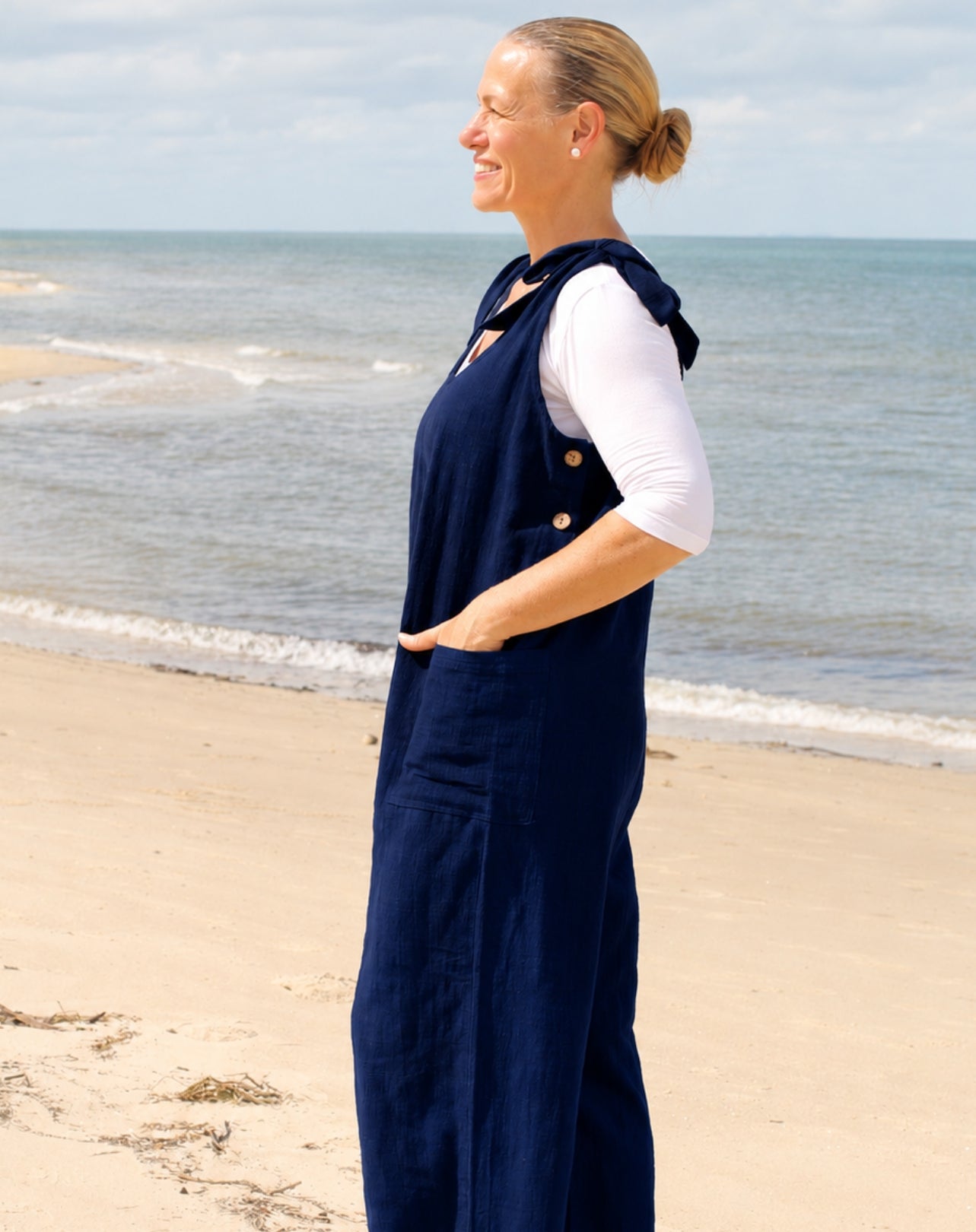 A woman standing on a sandy beach in profile, wearing a navy blue overall-style jumpsuit over a white long-sleeve top with functional buttons on side.
