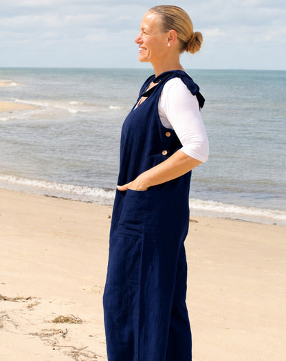 A woman standing on a sandy beach in profile, wearing a navy blue overall-style jumpsuit over a white long-sleeve top with functional buttons on side.