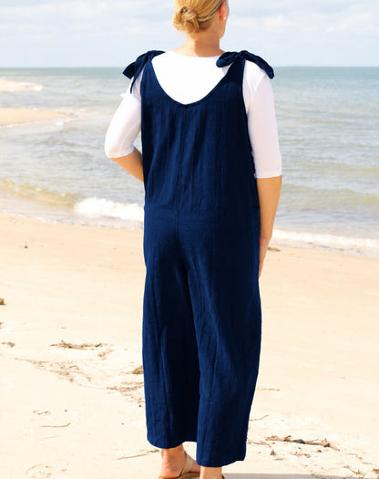 A woman standing on a sandy beach facing the ocean, wearing a navy blue wide-leg jumpsuit with tie straps over a white short-sleeve top.