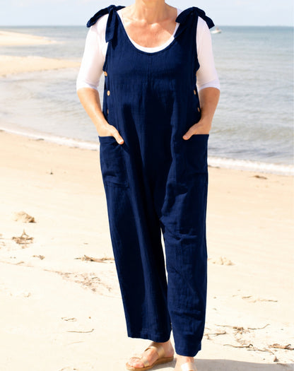 A woman wearing a navy blue linen-look overall jumpsuit with tie-shoulder straps and front pockets, layered over a white short-sleeve top, standing on a sandy beach with the ocean in the background.