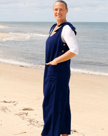 A woman standing on a sandy beach wearing a navy blue linen jumpsuit over a white short-sleeve top.