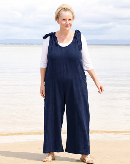 A woman wearing a navy blue wide-leg jumpsuit over a white short-sleeve top, standing on a sandy beach with water and a cloudy sky in the background.