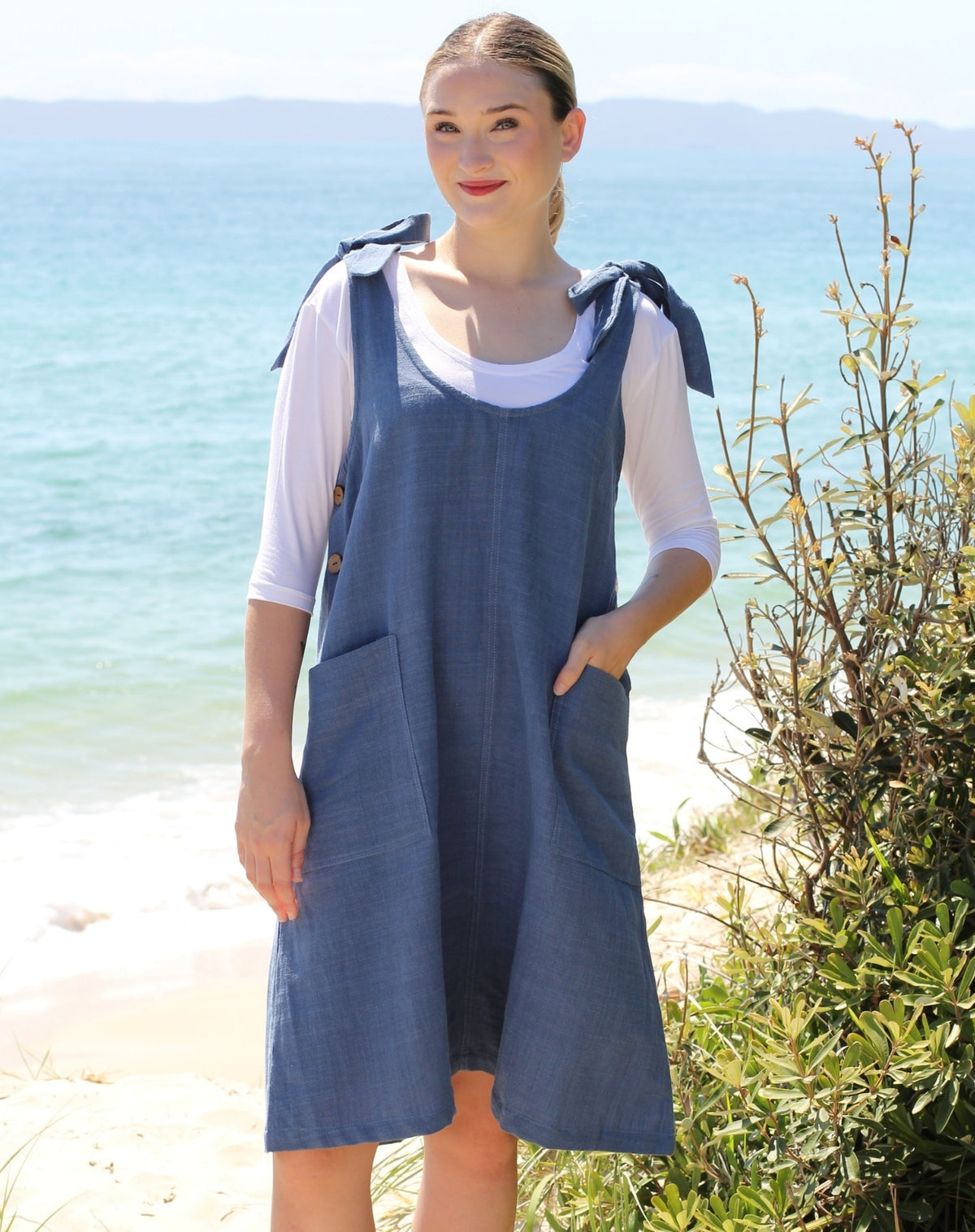 A woman standing on a beach wearing a blue denim pinafore dress over a white short-sleeve top. The dress has tie details on the shoulders and front patch pockets.