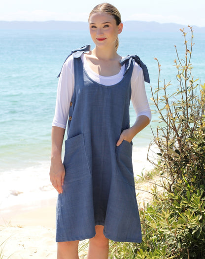 A woman standing on a beach wearing a blue denim pinafore dress over a white short-sleeve top. The dress has tie details on the shoulders and front patch pockets.
