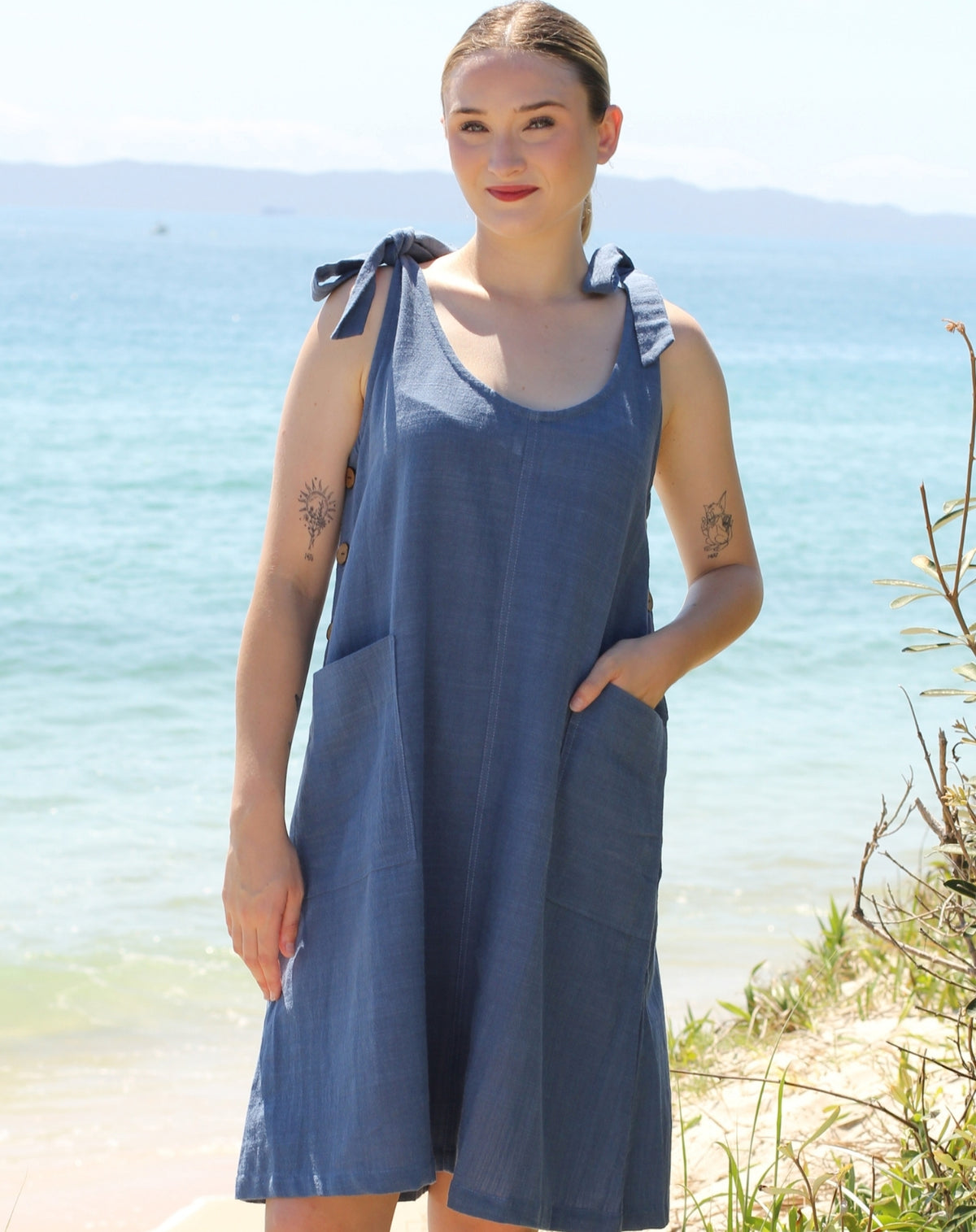 A woman wearing a sleeveless blue linen dress with tie-shoulder straps and front pockets, standing on a beach with the ocean and distant hills in the background.