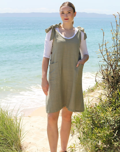 A woman wearing a sage green linen pinafore dress over a white t-shirt, standing on a sandy beach path with the ocean and greenery in the background.