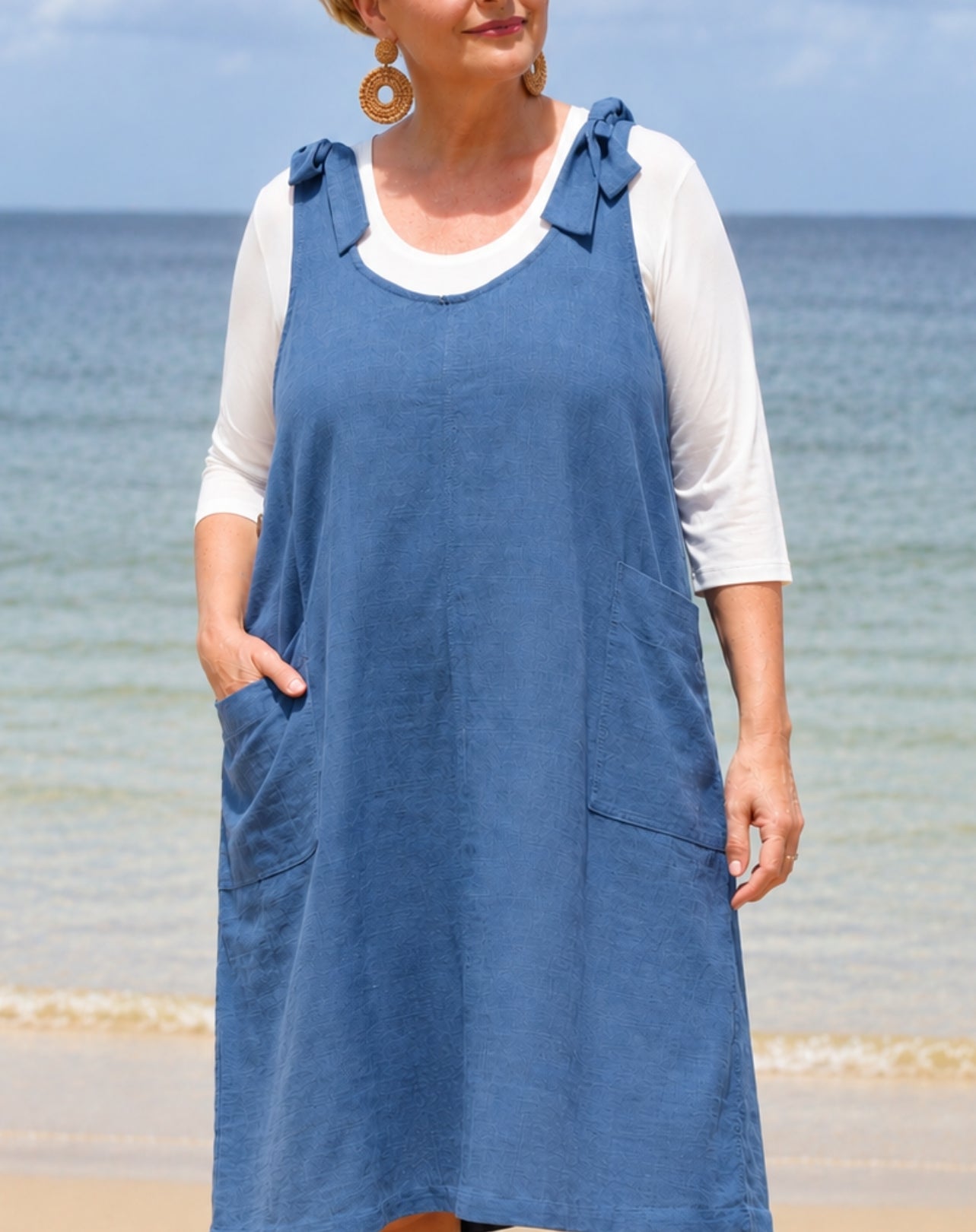 A woman wearing a blue linen cotton dress over a white 3/4-sleeve top, standing on a sandy beach with the ocean and sky in the background.