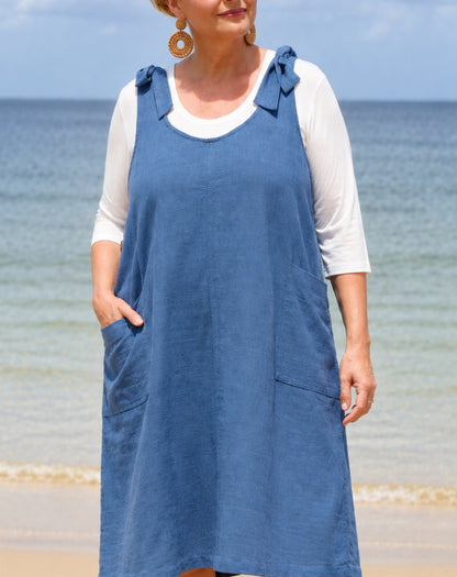 A woman wearing a blue linen cotton dress over a white 3/4-sleeve top, standing on a sandy beach with the ocean and sky in the background.