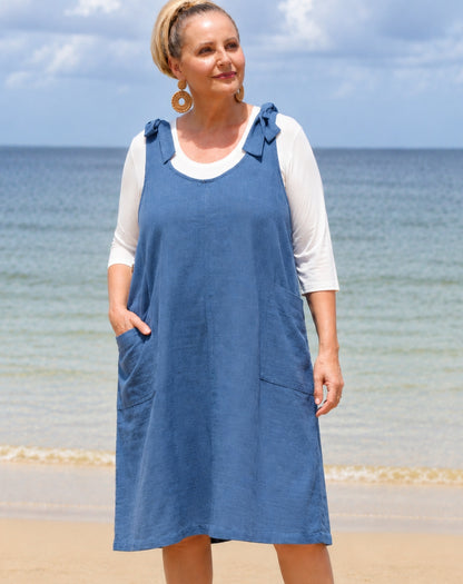 A woman wearing a blue linen pinafore dress over a white 3/4-sleeve top, standing on a sandy beach with the ocean and cloudy sky in the background.