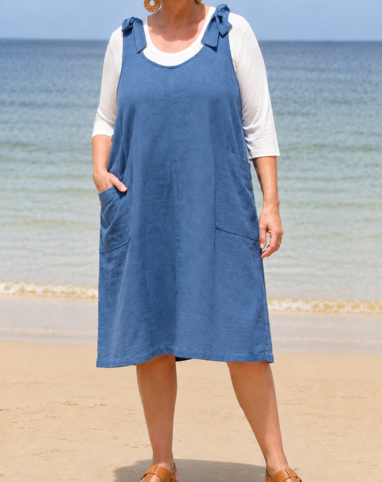 A woman wearing a blue cotton linen smock dress over a white 3/4 sleeve top, standing on a sandy beach with the ocean in the background.