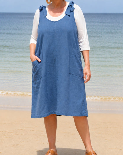 A woman wearing a blue cotton linen smock dress over a white 3/4 sleeve top, standing on a sandy beach with the ocean in the background.