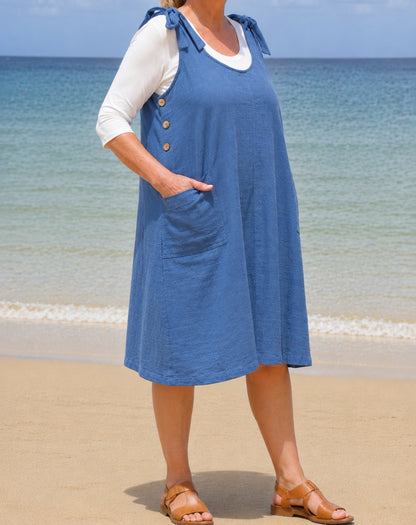 A woman wearing a blue sleeveless smock-style dress over a white long-sleeve top, standing on a sandy beach with the ocean in the background.