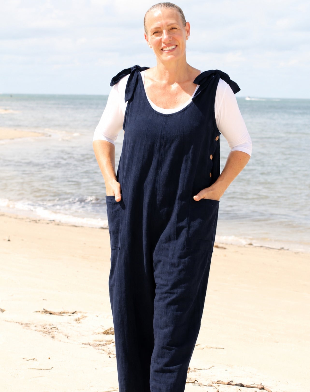 A woman standing on a sandy beach wearing a navy blue linen overall jumpsuit  with adjustable tie straps over a white long-sleeve t-shirt.