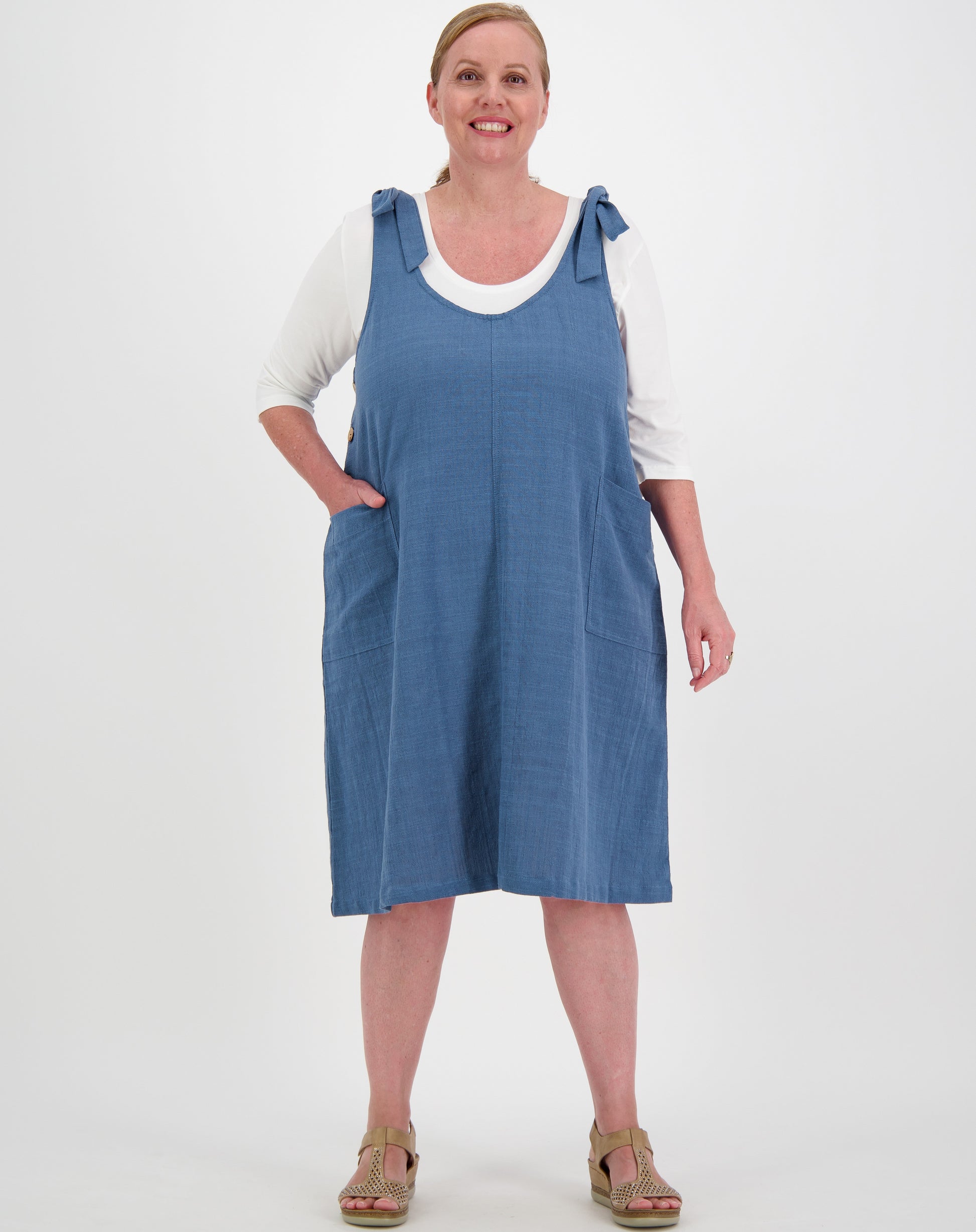 Woman wearing a blue denim tie strap smock dress over a white shirt on a white background