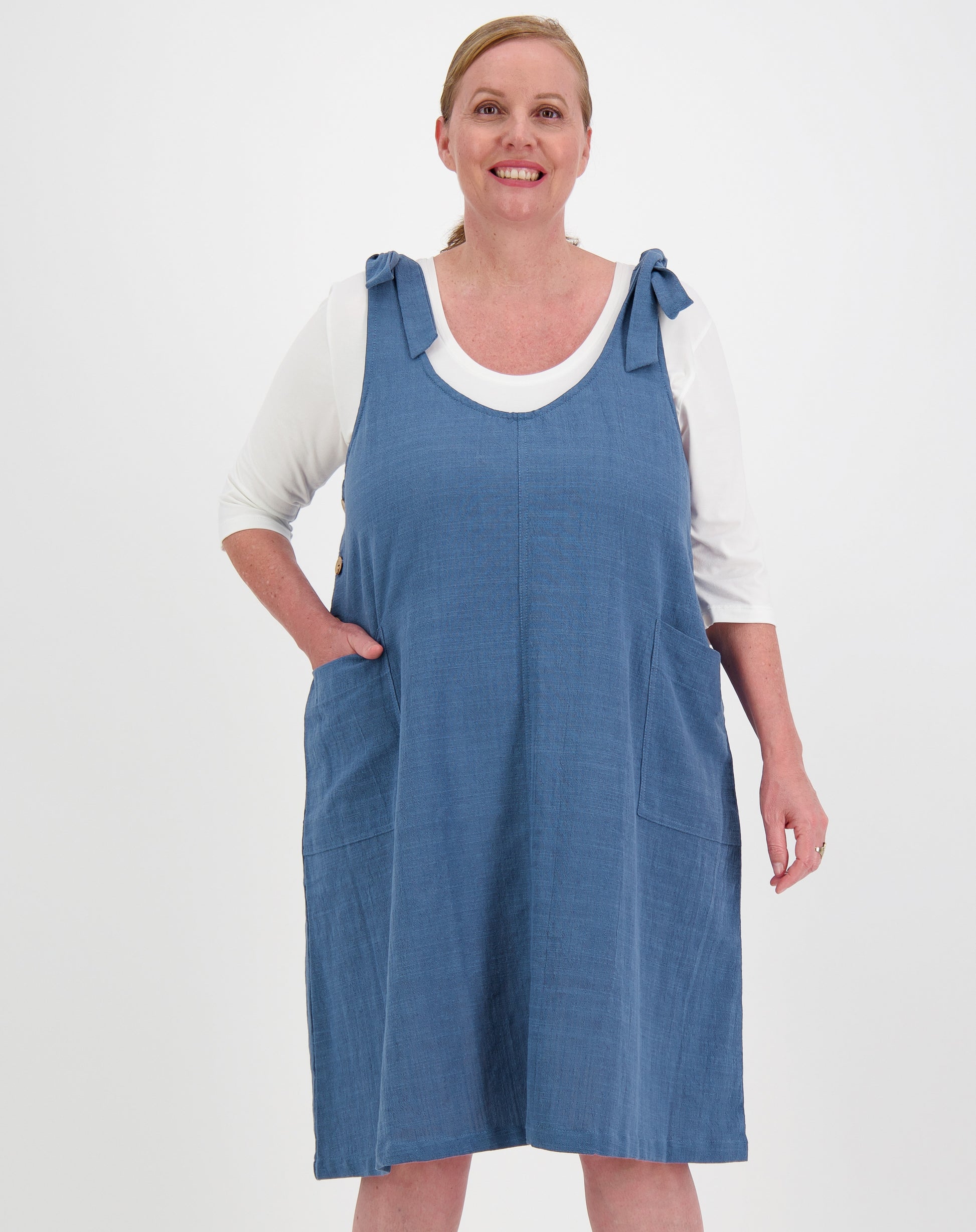 Woman wearing a blue tiestrap smock dress over a white shirt on a white background