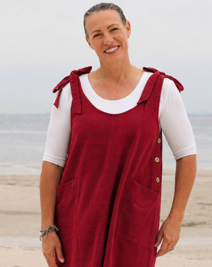 A smiling woman standing on a beach wearing a red linen overall dress over a white short-sleeve t-shirt. The dress features tie straps on the shoulders, button details on the side, and large front patch pockets.