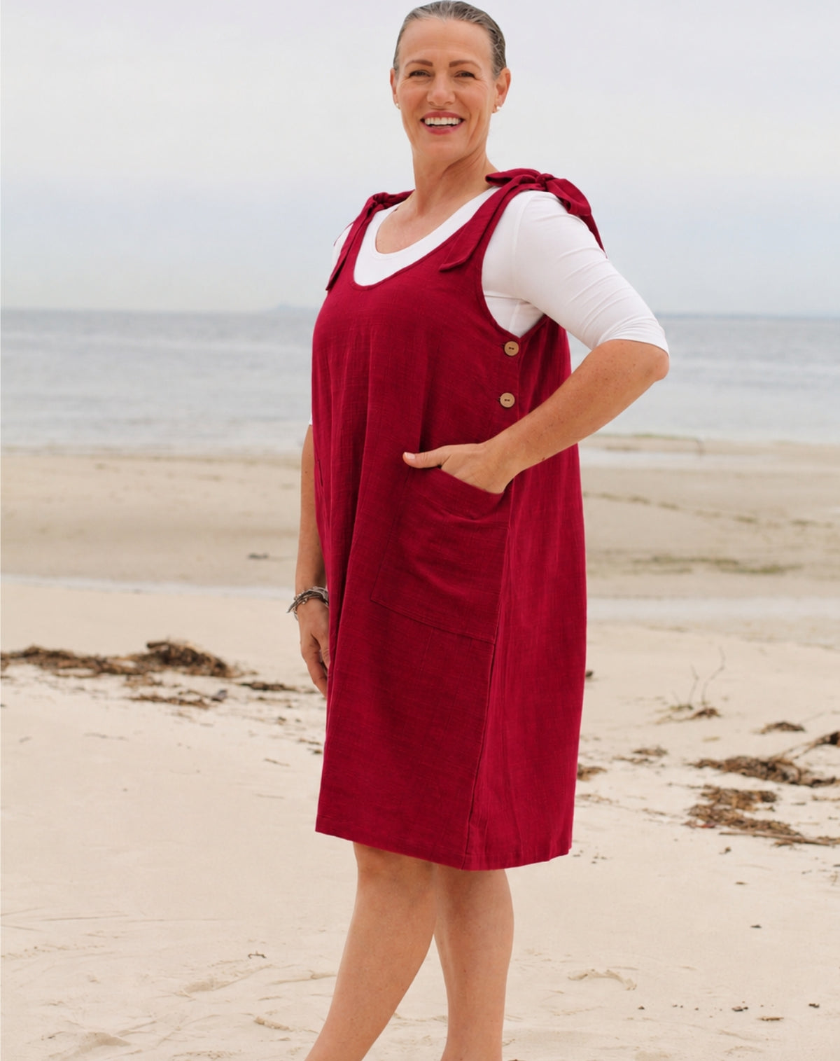 A woman standing on a sandy beach wearing a red linen look pinafore dress over a white short-sleeve top. The dress has a buttoned neckline and front pockets.