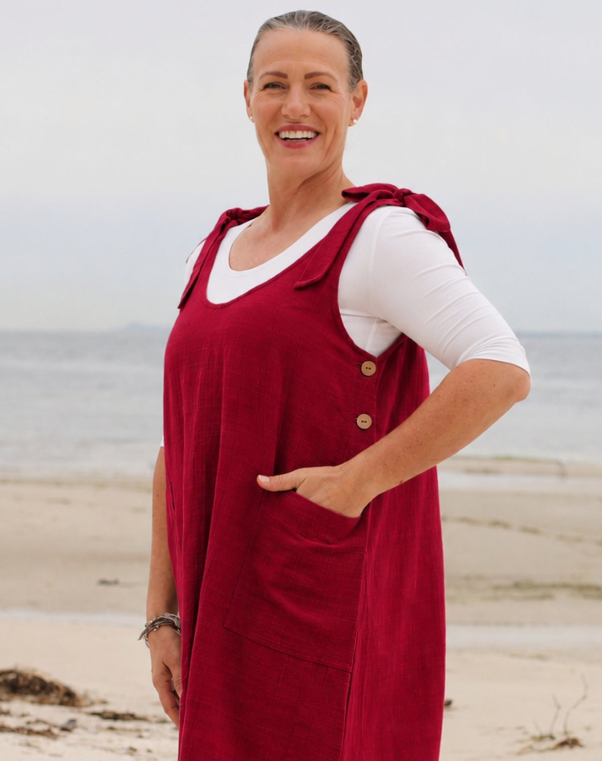 A woman standing on a beach wearing a red linen pinafore dress over a white long-sleeve t-shirt. The dress features a buttoned front, shoulder straps, and a large front pocket.