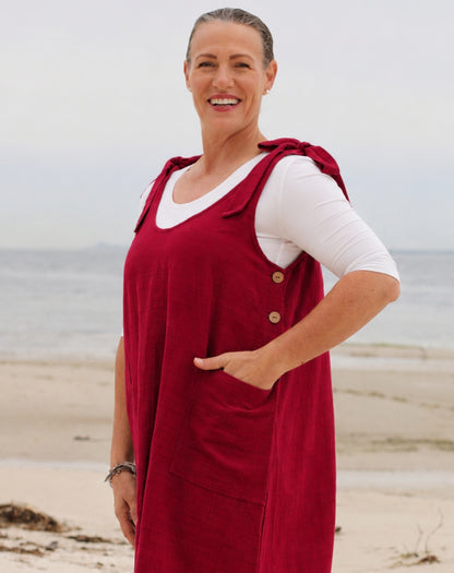 A woman standing on a beach wearing a red linen pinafore dress over a white long-sleeve t-shirt. The dress features a buttoned front, shoulder straps, and a large front pocket.