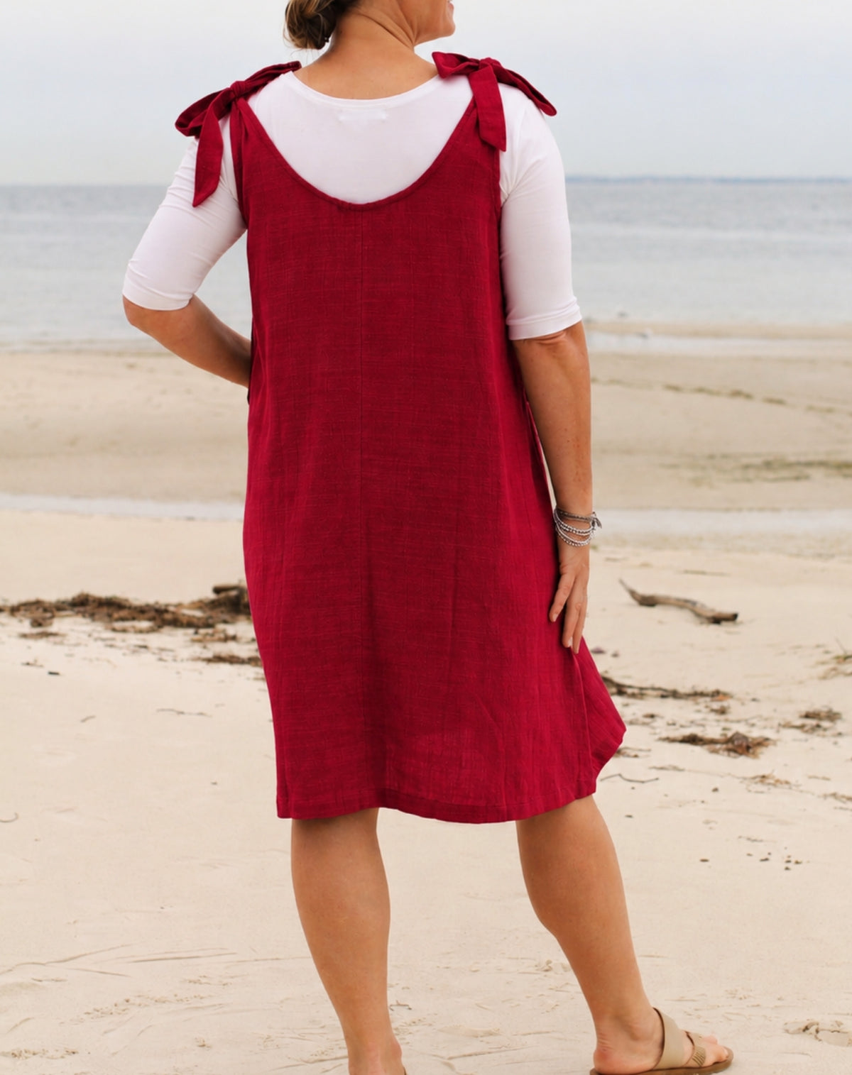 A woman standing on a sandy beach wearing a red sleeveless linen-look pinafore dress over a white short-sleeve t-shirt.