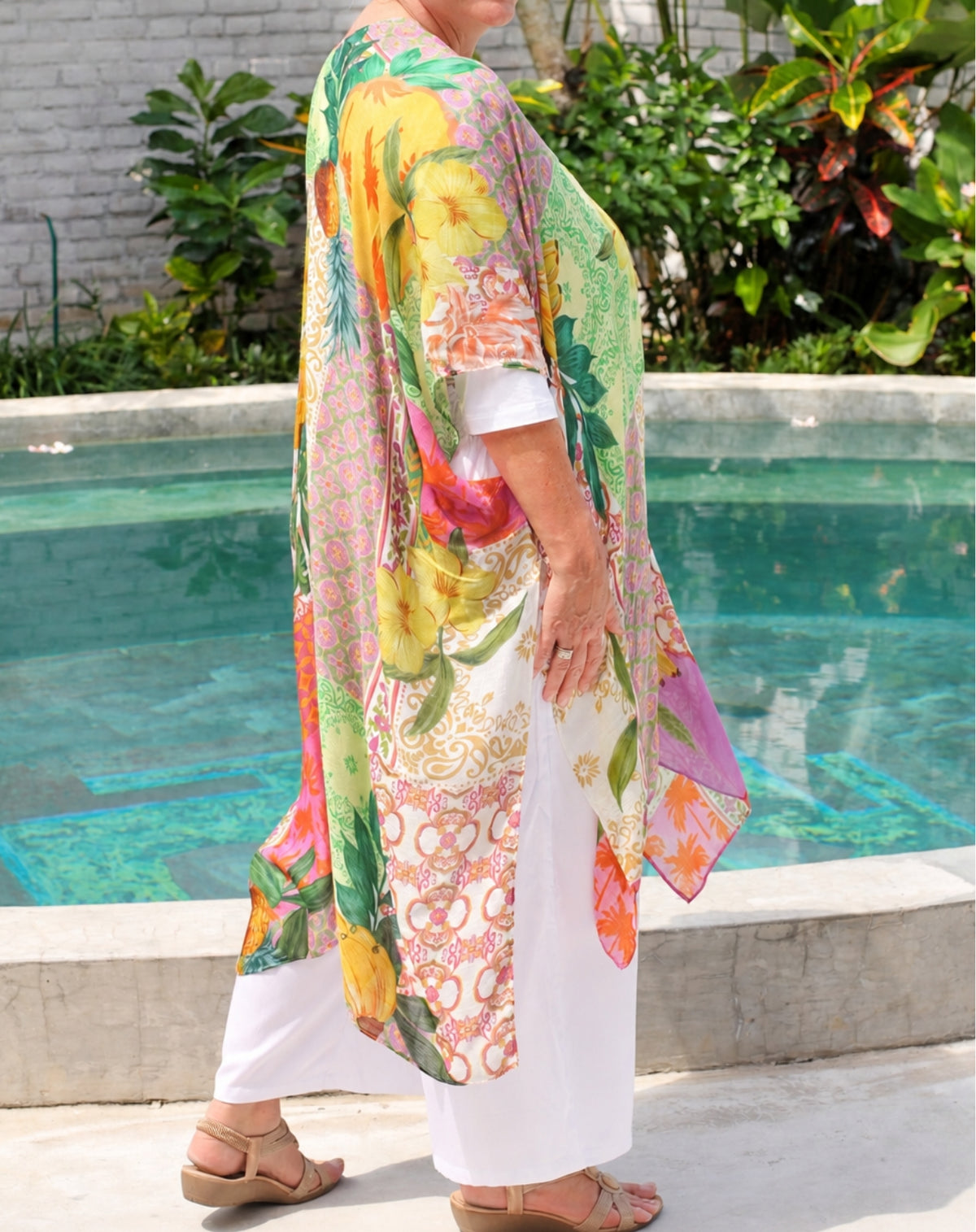 A woman wearing a colorful floral and paisley print kaftan tunic over white pants, standing next to a swimming pool in a garden setting.