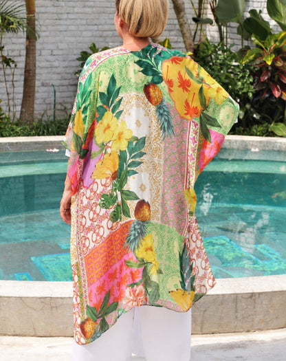A woman viewed from behind wearing a vibrant, multicolored tropical print kaftan or kimono with pineapples, hibiscus flowers, and lace patterns, standing by a swimming pool.