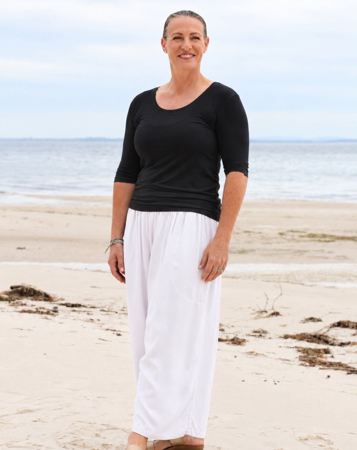 A woman standing on a sandy beach wearing a black 3/4-sleeve scoop-neck top and white wide-leg pants.
