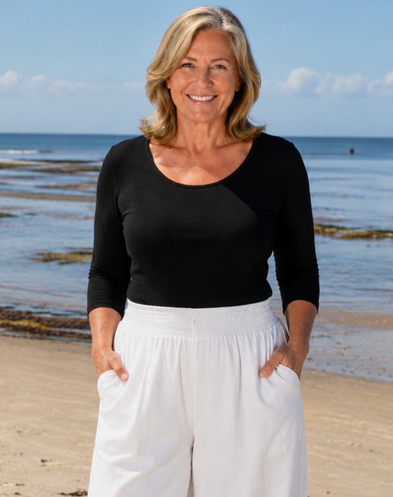 A smiling woman standing on a sandy beach wearing a black three-quarter-sleeve scoop-neck top and white pants, with the ocean and blue sky behind her.