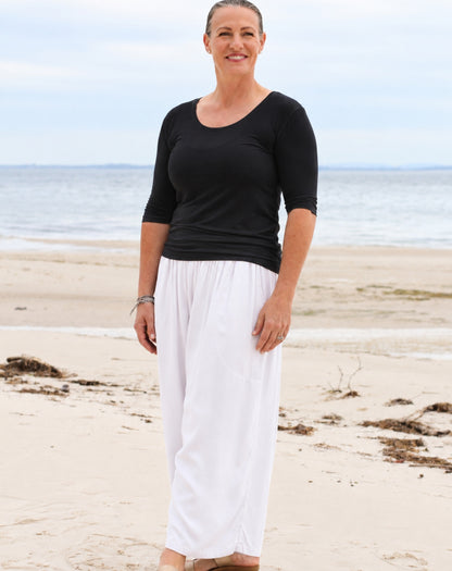 A woman standing on a sandy beach wearing a black three-quarter sleeve top and white wide-leg pants, with the ocean and cloudy sky in the background.