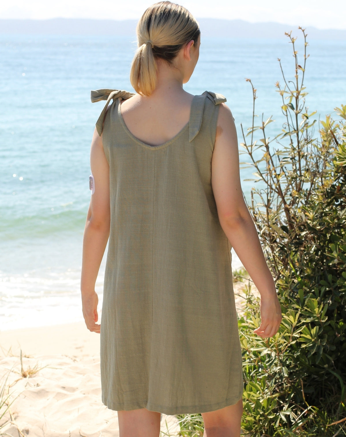 A woman viewed from behind wearing a sleeveless sage green linen smock dress with tie details on the shoulders, standing on a sandy beach with the ocean and greenery in the background.