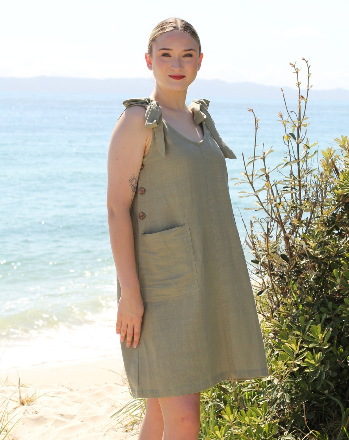 A woman wearing a sage green sleeveless tie strap smock dress with large front pocket, standing on a sandy beach with the ocean and greenery in the background.