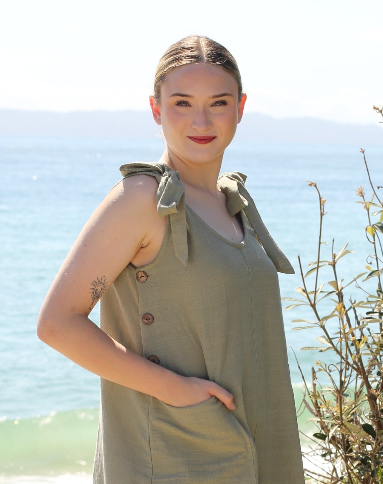 A woman wearing a sage green sleeveless smock dress with shoulder ties and front buttons, standing outdoors with the ocean and distant mountains in the background.