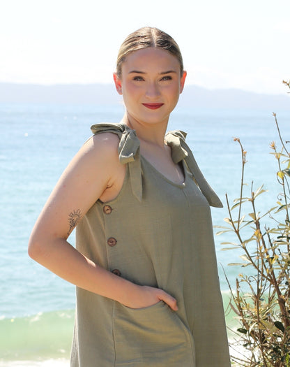 A woman wearing a sage green sleeveless smock dress with shoulder ties and front buttons, standing outdoors with the ocean and distant mountains in the background.